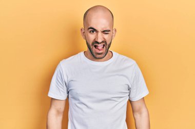 Gündelik beyaz tişört giyen genç kel adam kameraya seksi bir ifadeyle bakıyor, neşeli ve mutlu bir yüzle.. 