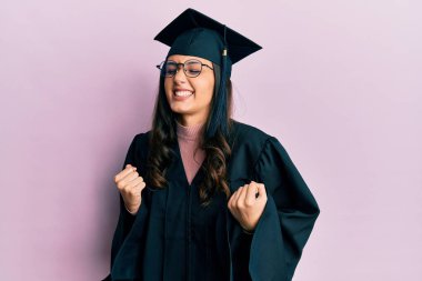 Mezuniyet şapkası ve tören cübbesi takan genç İspanyol kadın çok mutlu ve heyecanlı bir şekilde kollarını kaldırıp, gülümseyerek ve başarı çığlıkları atarak zafer jesti yapıyor. kutlama konsepti. 