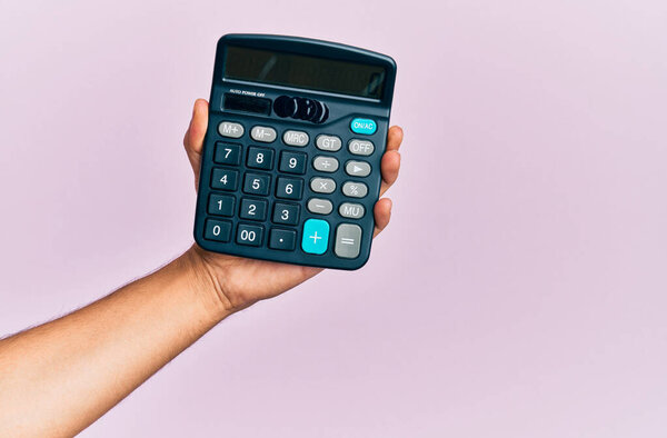 Hand of young hispanic man holding calculator over isolated pink background.