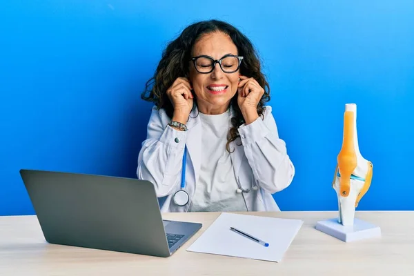 Beautiful middle age woman doctor at orthopedic clinic covering ears with fingers with annoyed expression for the noise of loud music. deaf concept.