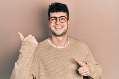 Gündelik kıyafetler giyen ve gözlüklü genç bir adam arka tarafı işaret ediyor. Elleri havada, kendinden emin bir şekilde gülümsüyor. 