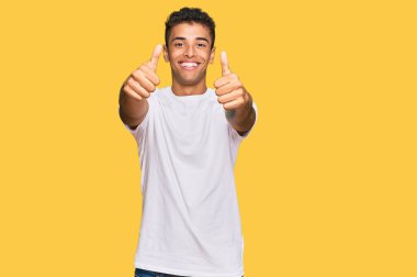 Genç ve yakışıklı Afro-Amerikan erkeği, günlük beyaz tişört giyiyor. Elle olumlu jest yapmayı onaylıyor. Gülümsüyor ve başarı için seviniyor. kazanan jest. 