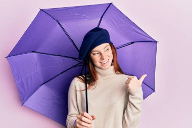 Mor şemsiyeli genç, kızıl saçlı kız taze bere takıyor. Başparmağıyla yan tarafa bakıyor. Mutlulukla gülümsüyor. 