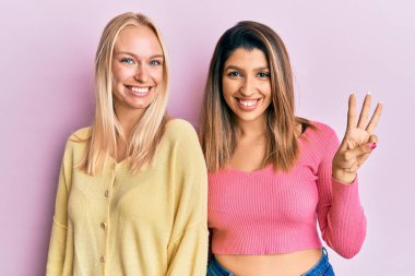 Pembe arka planda yan yana duran iki arkadaş üç numaralı parmaklarını gösterip işaret ederken kendinden emin ve mutlu gülümsüyor.. 
