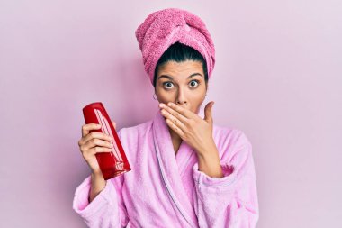 Duş bornozu giymiş, ağzı şampuanla kaplı, ağzı elle kaplanmış, şok olmuş ve hata yapmaktan korkan genç bir İspanyol kadın. şaşırmış ifade 