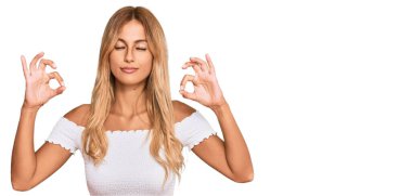 Güzel sarışın genç kadın, günlük beyaz tişört giyip rahatlıyor ve gözleri kapalı gülümsüyor parmaklarıyla meditasyon hareketi yapıyor. yoga konsepti. 