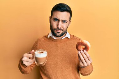 Genç İspanyol adam donut yiyor ve kahve içiyor şüpheci ve gergin, sorun yüzünden surat asıyor. negatif kişi. 