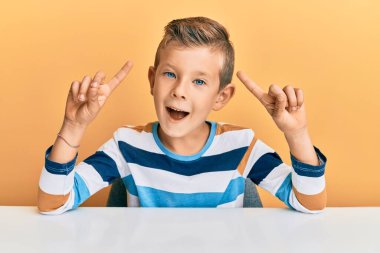 Sevimli beyaz çocuk, günlük giysiler içinde masada oturmuş şaşkın şaşkın şaşkın gülüyor, parmaklarıyla işaret ediyor ve kollarını kaldırıyordu.. 