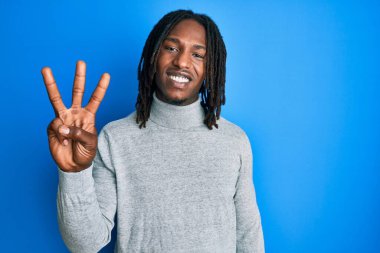 Örgülü, balıkçı yaka kazaklı Afro-Amerikalı bir adam üç numaralı parmaklarıyla gülümseyerek, kendinden emin ve mutlu bir şekilde öne çıkıyor.. 