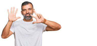 Orta yaşlı, beyaz tişört giyen yakışıklı bir adam 8 numaralı parmaklarıyla gösterip işaret ederken kendinden emin ve mutlu gülümsüyor.. 