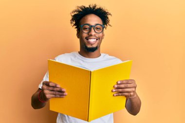 Sakallı, kitap okuyan, gözlüklü mutlu ve havalı bir gülümsemeyle gülümseyen genç bir Afrikalı Amerikalı. diş gösterme. 