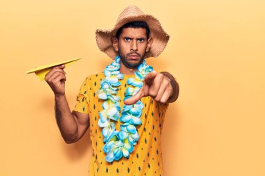 Yaz şapkası takan genç bir latin ve elinde kağıt uçakla kamerayı ve seni işaret eden Hawaii zambağı tutan kendine güvenen bir jest. 