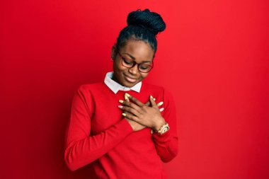 Sıradan kıyafetler ve gözlük takan, elleri göğsünde, gözleri kapalı gülümseyen ve yüzünde minnet ifadesi olan Afro-Amerikan genç bir kadın. sağlık kavramı. 