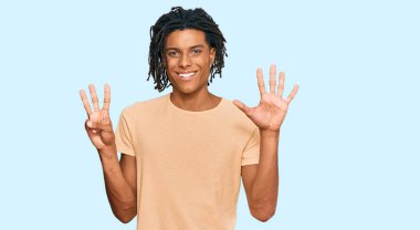 Genç, Afro-Amerikalı, günlük giysiler giymiş, 8 numaralı parmaklarıyla işaret ederken, kendinden emin ve mutlu bir şekilde gülümsüyor.. 