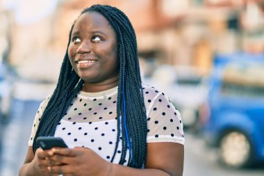 Genç Afrikalı Amerikalı kadın şehirde akıllı telefon kullanarak mutlu bir şekilde gülümsüyor..