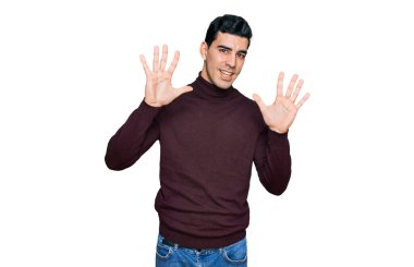 Yakışıklı, balıkçı yaka kazak giyen, kendini beğenmiş ve mutlu bir şekilde gülümseyen, on numarayı gösteren İspanyol bir adam.. 