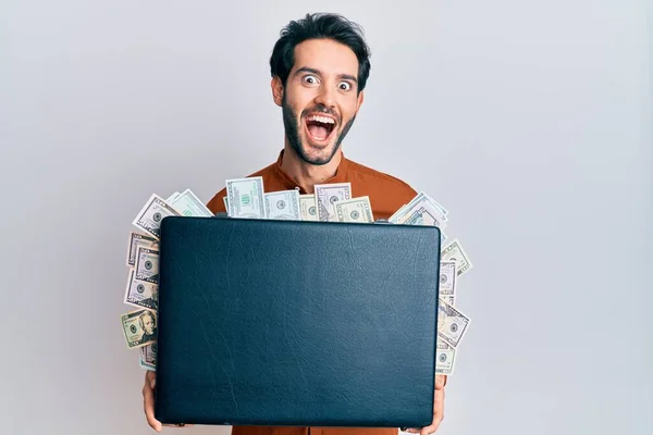 Young hispanic man holding briefcase with dollars celebrating crazy and amazed for success with open eyes screaming excited.