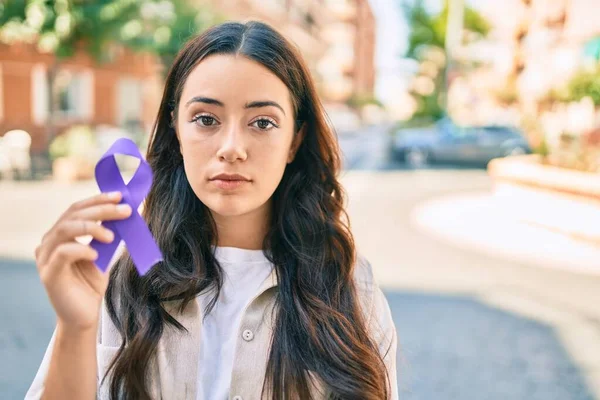 Young hispanic woman with serious expression holding purple ribbon walking at the city.