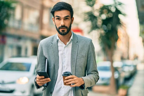 Young hispanic businessman with serious expression using smartphone and drinking coffee at the city.