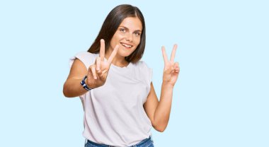 Beyaz bir tişört giyen genç beyaz kadın kameraya bakıp zafer işareti yapan parmaklarını gösteriyor. İki numara.. 