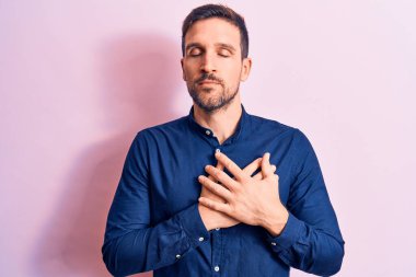 Genç ve yakışıklı bir adam pembe arka plan üzerinde dikiliyor. Elleri göğsünde, gözleri kapalı ve yüzünde minnet dolu bir jest ile gülümsüyor. Sağlık kavramı.