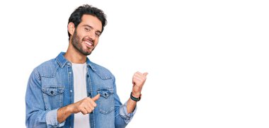 Gündelik kıyafetler giyen genç İspanyol adam arka tarafı işaret ediyor. Elleri havada, kendinden emin bir şekilde gülümsüyor. 