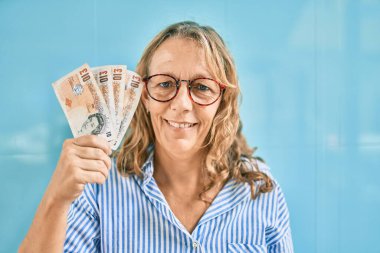 Orta yaşlı, beyaz bir kadın, elinde İngiliz banknotlarıyla şehirde mutlu bir şekilde gülümsüyor..