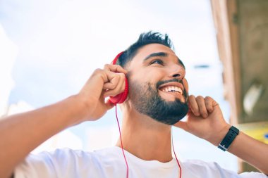 Genç Arap adam şehirde yürürken kulaklık takarak mutlu bir şekilde gülümsüyor..