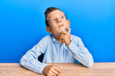 Kafkasyalı sevimli bir çocuk, günlük giysiler içinde masaya oturmuş şüphe hakkında kafa yoruyor, çenesi parmaklarıyla yukarı bakıyor ve merak ediyor. 