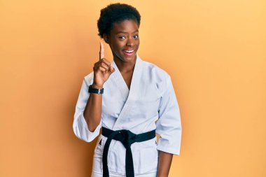 Genç Afro-Amerikalı kız karate kimonosu ve siyah kuşak giyiyor. Bir numarayı işaret ediyor ve gülümsüyor. Kendinden emin ve mutlu.. 