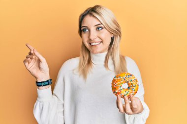 Genç beyaz kadın elinde donut tutarken mutlu bir şekilde gülümsüyor. 
