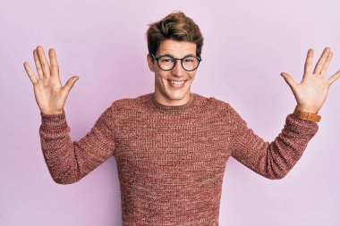 Yakışıklı, beyaz bir adam günlük süveter giyiyor ve gözlük takıyor. 10 numaralı parmaklarıyla işaret ediyor. Kendinden emin ve mutlu bir şekilde gülümsüyor.. 