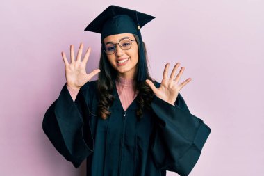 Mezuniyet şapkası ve tören cübbesi takan genç İspanyol kadın on numarayı gösterip işaret ederken kendinden emin ve mutlu gülümsüyor.. 