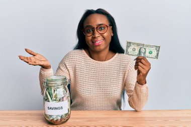 Genç Afrikalı bir kadın elinde dolar ve kavanozla zaferi kutluyor mutlu bir gülümsemeyle ve elini kaldırarak kazanan bir ifadeyle. 