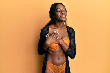 Genç Afro-Amerikalı kadın iç çamaşırları giyiyor. Elleri göğsünde, gözleri kapalı gülümsüyor ve yüzünde minnet ifadesi var. sağlık kavramı. 