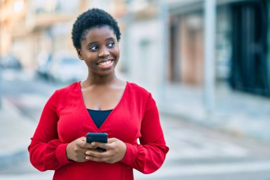 Genç Afrikalı Amerikalı kadın şehirde akıllı telefon kullanarak mutlu bir şekilde gülümsüyor..