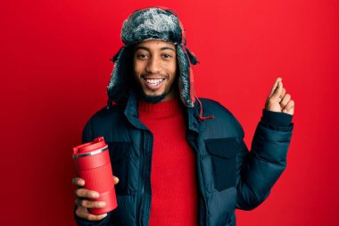 Sakallı, kışlık şapkalı, termosu üstünde kahve tutan, gülümseyen, mutlu, eli ve parmağı kenarda genç bir Afrikalı Amerikalı. 