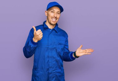 Kel, sakallı, inşaatçı tulumu giyen, avuç içi gösteren ve baş parmağıyla hareket eden mutlu ve neşeli gülümseyen bir adam. 