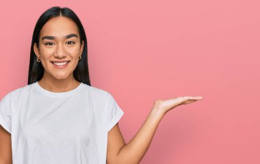 Genç Asyalı kadın, günlük beyaz t-shirt giyip neşeli bir şekilde gülümsüyor ve avuç içiyle kameraya bakıyor.. 