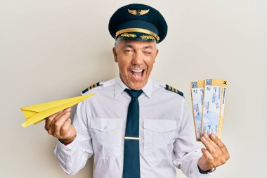 Yakışıklı orta yaşlı yetişkin pilot elinde kağıt uçak ve geçiş kartı ile kameraya seksi bir ifadeyle bakıyor, neşeli ve mutlu bir yüzle.. 