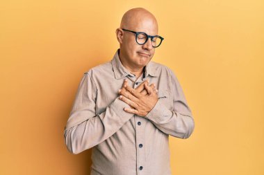 Orta yaşlı kel bir adam günlük kıyafetler ve gözlük takıyor elleri göğsünde, gözleri kapalı gülümsüyor ve yüzünde minnet dolu bir jest var. sağlık kavramı. 