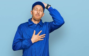 Sakallı kel bir adam. Üniforma giyiyor. Hastalık ve ateş için alnına dokunuyor. 