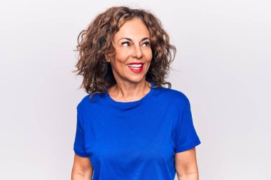 Orta yaşlı, güzel, esmer, mavi tişörtlü bir kadın beyaz arka plan üzerinde duruyor, yan tarafa bakıyor, rahat bir görünüm takınıyor ve kendine güvenen bir gülümseme sergiliyor..