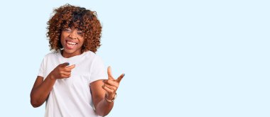 Gündelik beyaz tişört giyen genç Afro-Amerikalı kadın mutlu ve komik yüzlü parmaklarını kameraya doğrultuyor. iyi enerji ve titreşimler. 