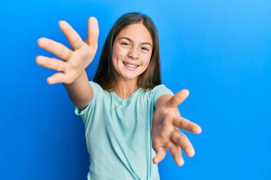 Güzel, esmer, beyaz tişört giyen küçük bir kız kameraya sarılıp gülümsüyor. Mutluluğu kucaklayan neşeli bir ifade.. 