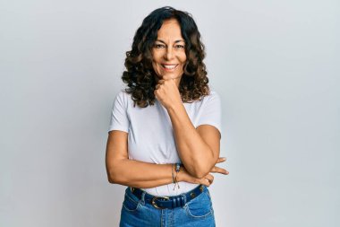 Orta yaşlı İspanyol kadın, günlük beyaz tişört giyiyor kameraya kendinden emin bakıyor kollarını kavuşturmuş ve çenesini kaldırmış gülümsüyor. Olumlu düşünmek. 