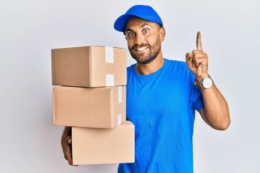 Sakallı, kurye üniformalı yakışıklı teslimat paketleri taşıyan, şaşkın, şaşkın, parmaklarıyla işaret eden ve kollarını kaldıran bir adam.. 