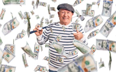 Golf sopası ve top tutan yaşlı, beyaz saçlı yakışıklı adam mutlu ve pozitif gülümsüyor, başparmağıyla mükemmel ve onay işareti yapıyor.