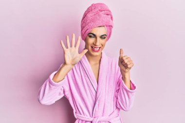 Kafasına duş havlusu ve bornoz takmış genç adam altı numaralı parmaklarını gösterip onu gösterirken kendinden emin ve mutlu bir şekilde gülümsüyor.. 