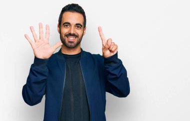 Latin asıllı genç adam gündelik giysiler giyip altı numaralı parmaklarıyla işaret ederken kendinden emin ve mutlu bir şekilde gülümsüyor.. 